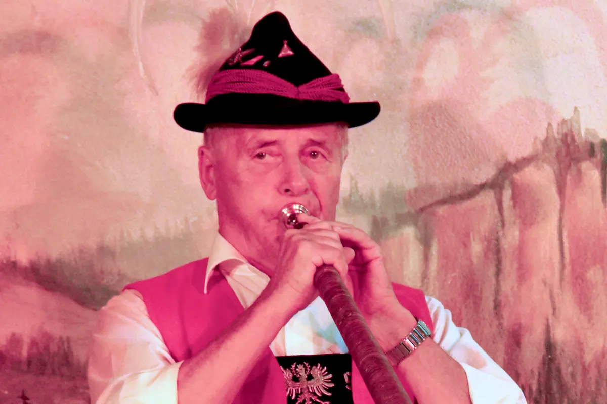 A man in a pink waistcoat and a hat with a tall feather blows a long alphorn in front of a painted cliff backdrop