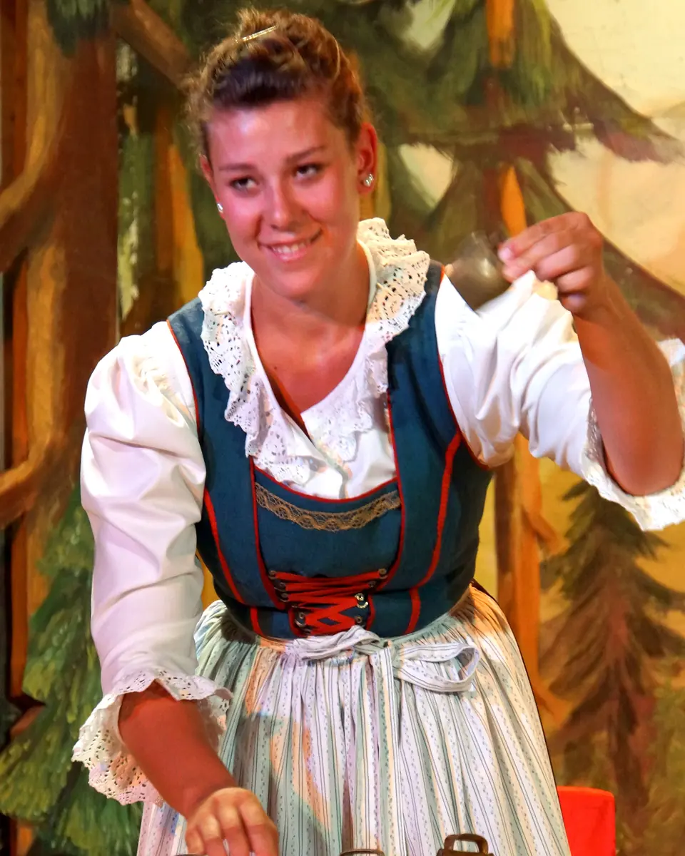 A woman in a dirndl rings a row of cowbells set on a red and white checked table in front of painted fir trees