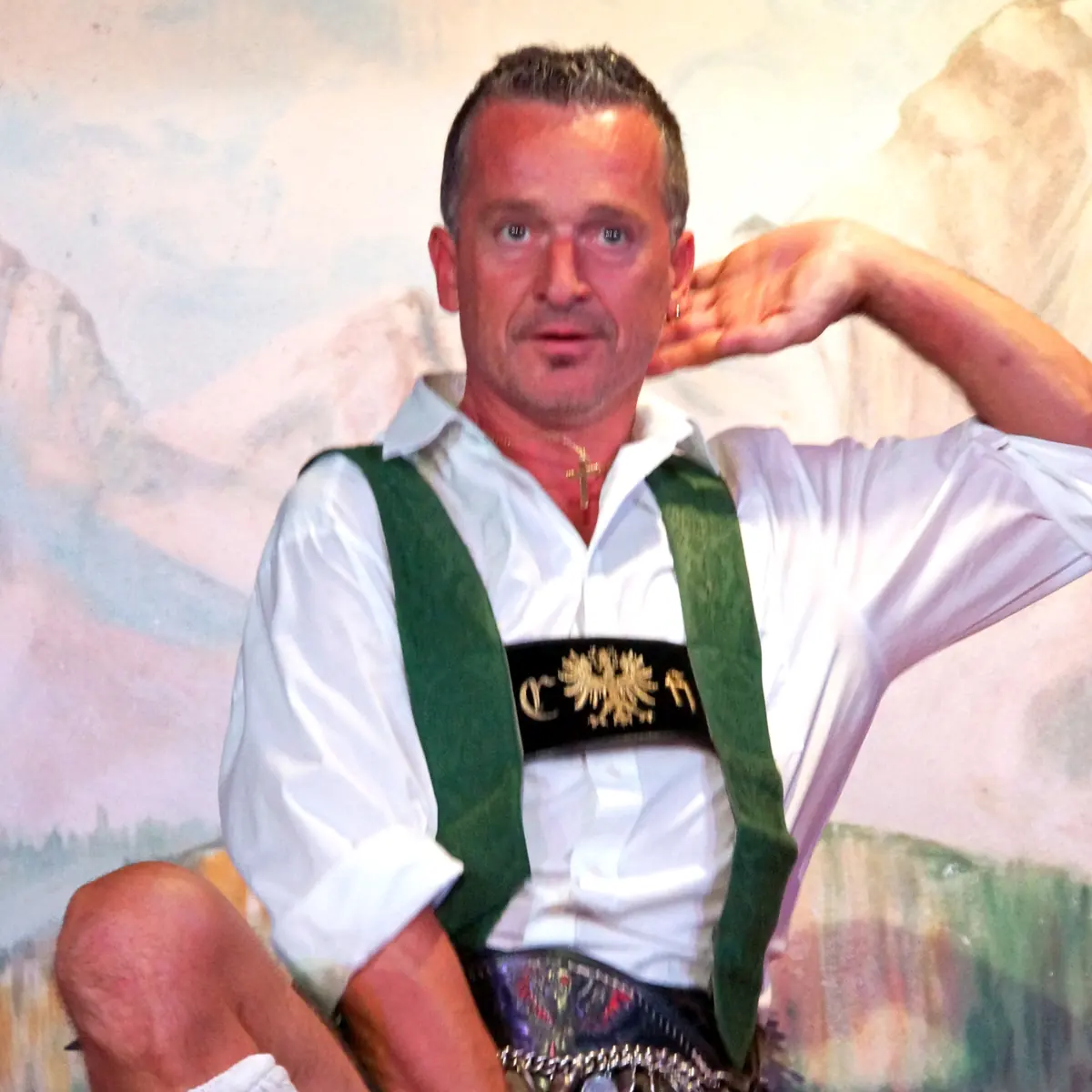 A man in lederhosen and green braces dances with one knee raised and a hand behind his head, in front of a painted mountain backdrop