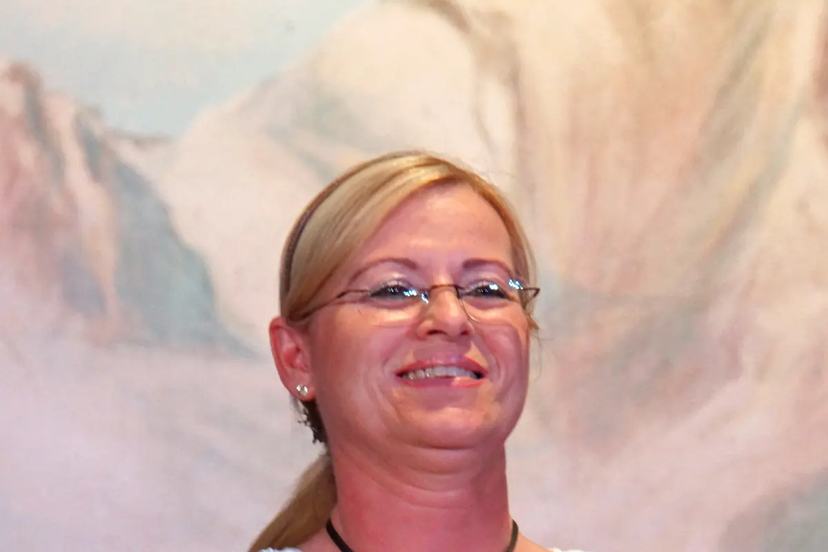 A smiling woman in glasses, a white blouse and a dark bodice stands in front of a painted mountain backdrop