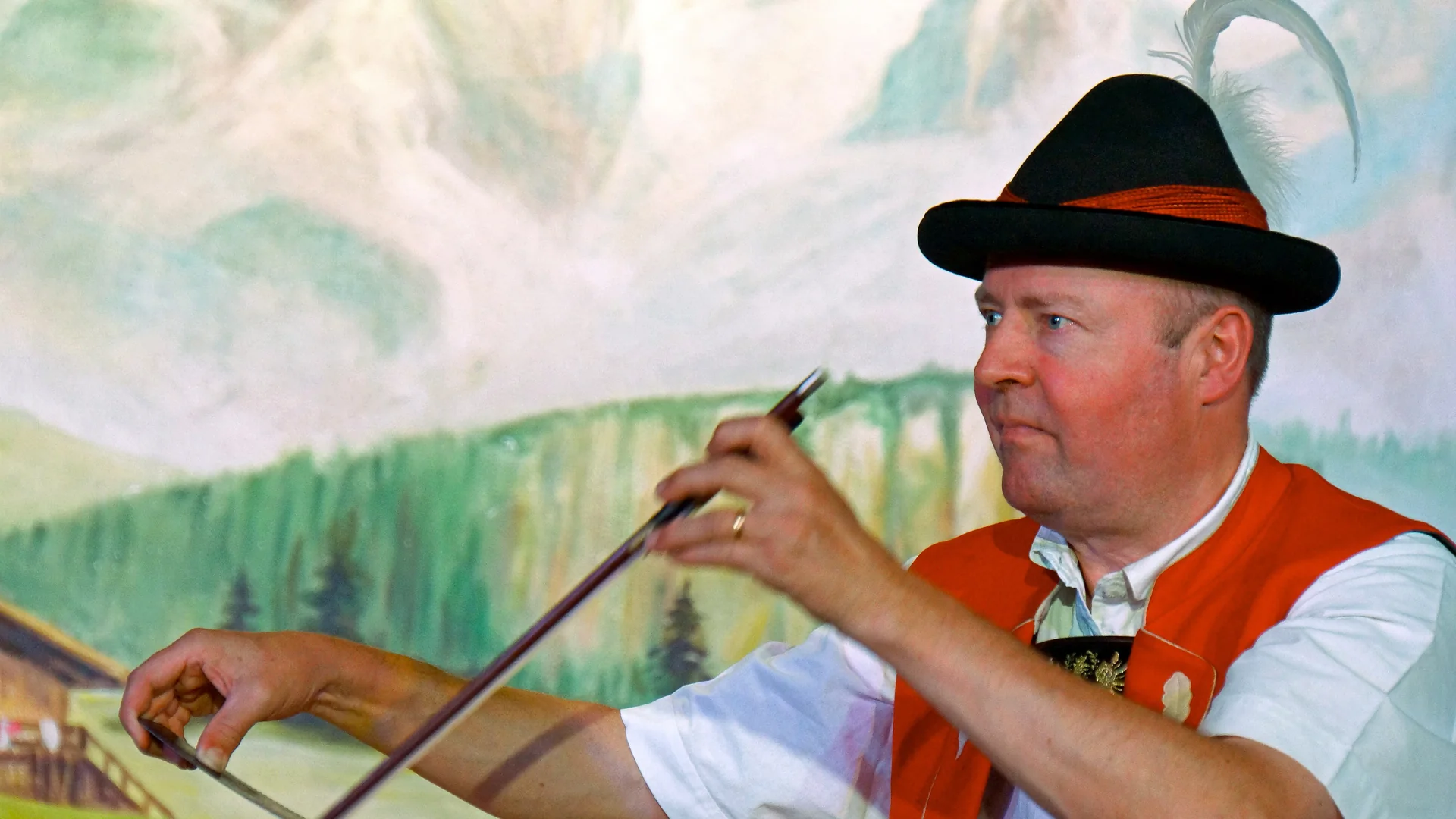 A musician in a red waistcoat and a feathered hat draws a bow across a singing saw in front of a painted alpine meadow and cliffs